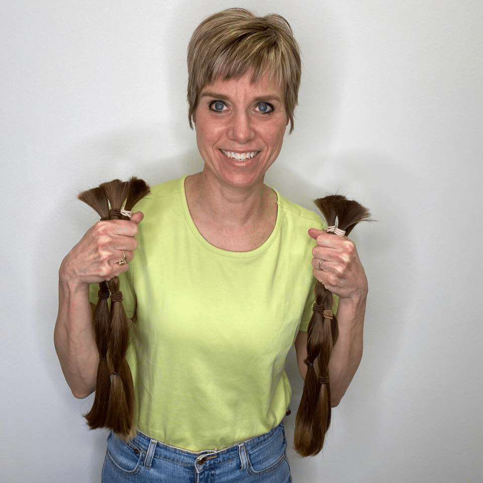 A white woman with a short dirty blonde pixie cut wearing a neon green tshirt and jeans. She is holding six, 16 inch-long ponytails of her own, brown hair.