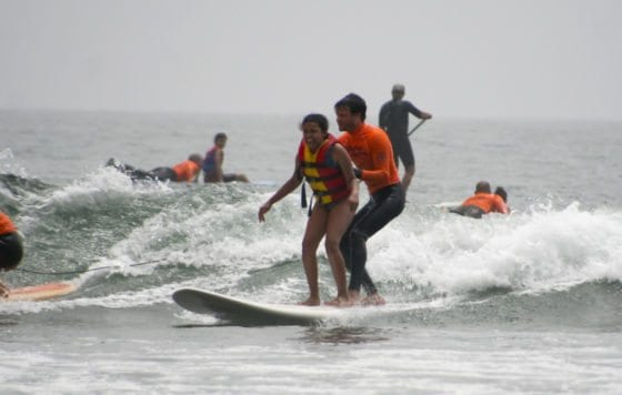 An adaptive surfing instructor and student stand on a surfboard together and ride a wave.